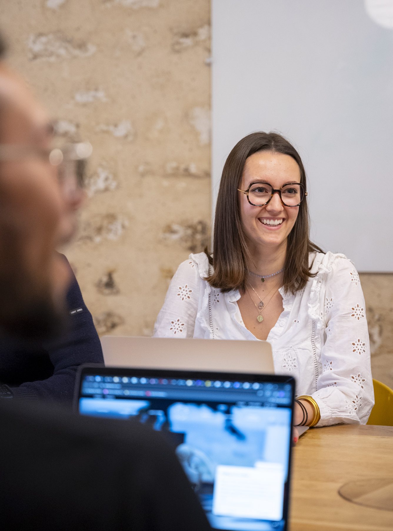 Une femme souriante portant une chemise blanche est assise à une table dans un environnement de bureau, avec un ordinateur portable visible au premier plan.