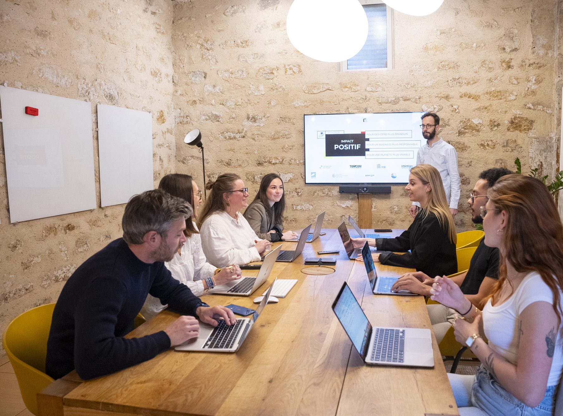 Une équipe de jeunes professionnels discute autour d'une table en bois, avec des ordinateurs portables ouverts, dans une salle de réunion moderne avec des murs en pierre.