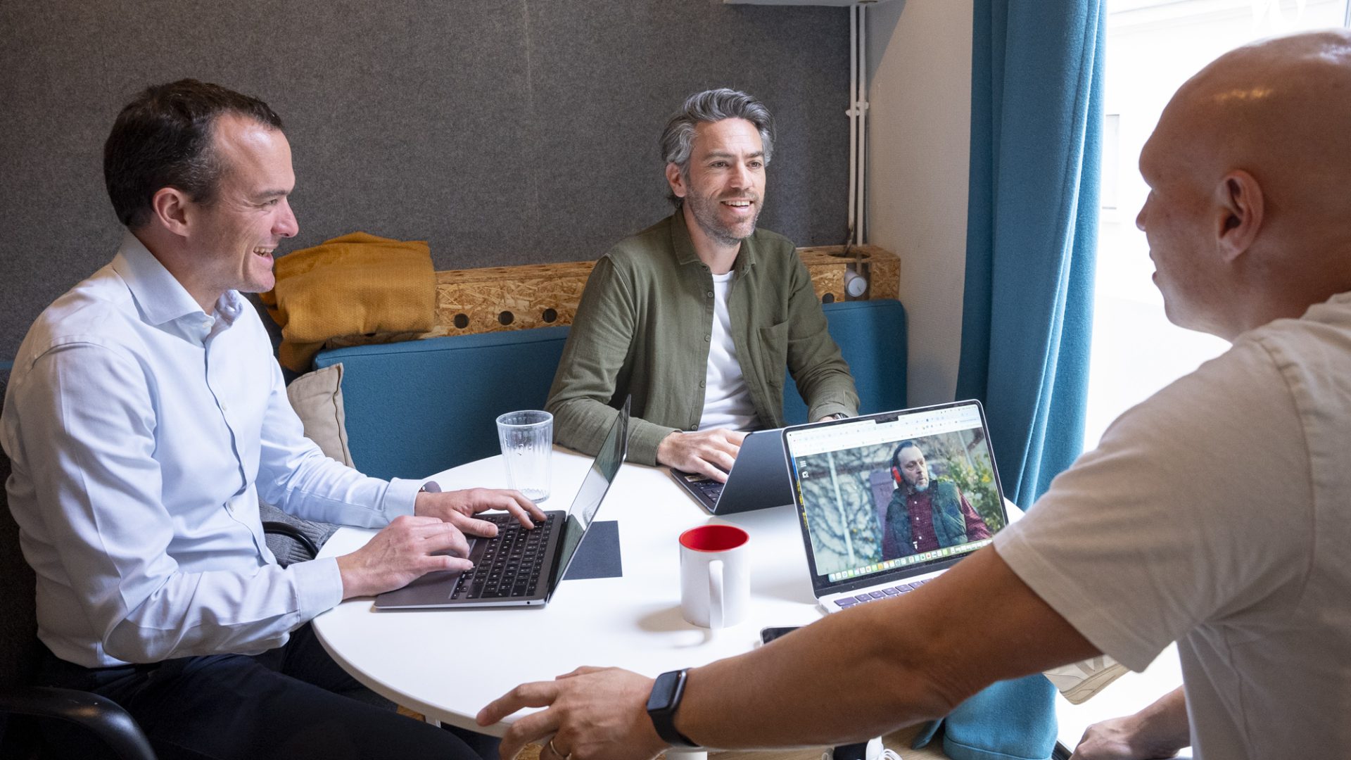 Trois hommes sont assis autour d'une table, travaillant sur des ordinateurs portables et discutant dans un espace de réunion lumineux.