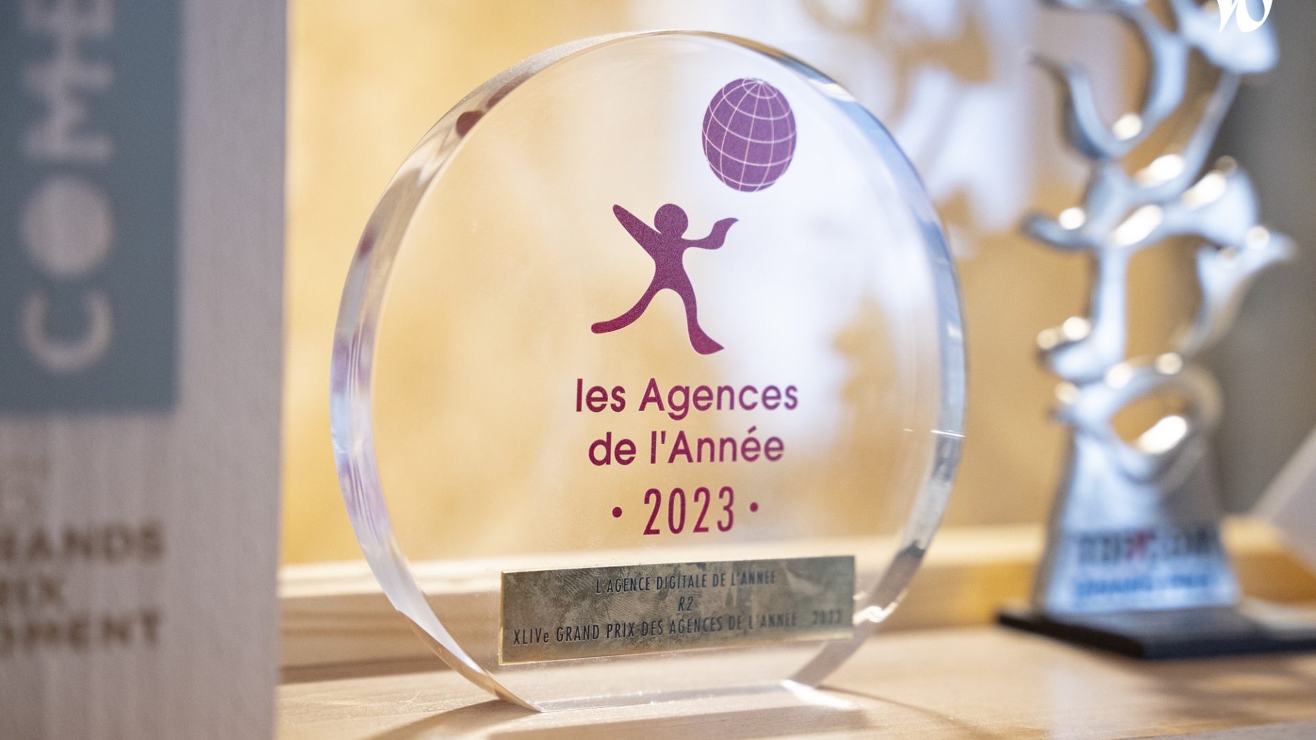 Un prix en verre gravé indiquant "les Agences de l’Année 2023" avec une mention spéciale pour "l’Agence Digitale de l’Année" à côté d'une autre statuette.
