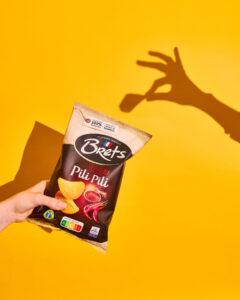 Un sachet de chips "Pili Pili" de la marque Brets est tenu devant un fond jaune vif, projetant l'ombre d'une main en forme de pincement.